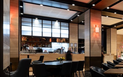 Dark-toned dining room with black leather chairs, round tables, wood-panelled walls, bar visible through opening