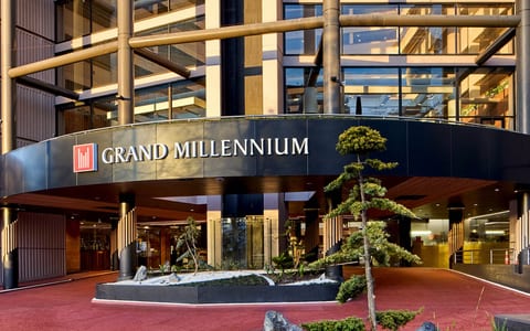 Grand Millennium hotel entrance with curved canopy, timber accents, and sculpted trees