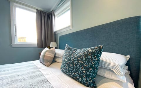 Blue-walled bedroom with upholstered headboard, layered cushions, and curtained window
