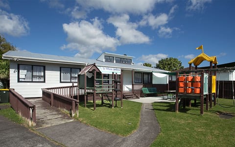 Single-storey white weatherboard community building with playground and garden