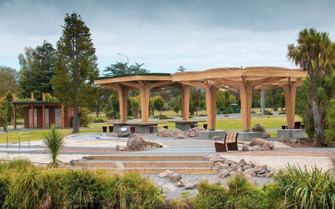 Timber open pavilion structure with latticed roof beams, concrete seating steps and boulders, parkland setting