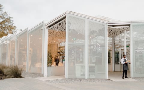 White glazed pavilion exterior with perforated roof panels, timber columns and gravel surrounds