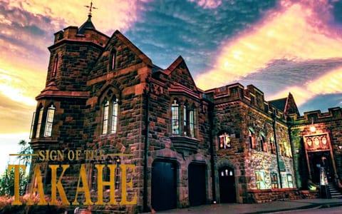 Gothic stone building with turrets, arched windows and a central tower, lit at dusk against a dramatic sky