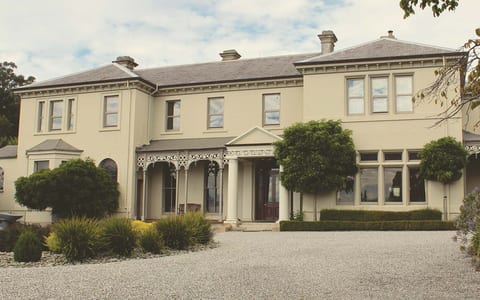 Two-storey cream Victorian manor with verandah and arched windows behind a gravel driveway and clipped hedges