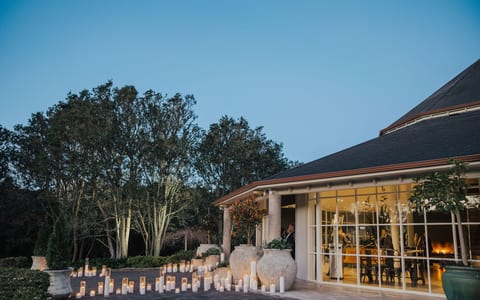 Low-lit exterior at dusk with rows of candles on a paved terrace, glazed pavilion and trees in background