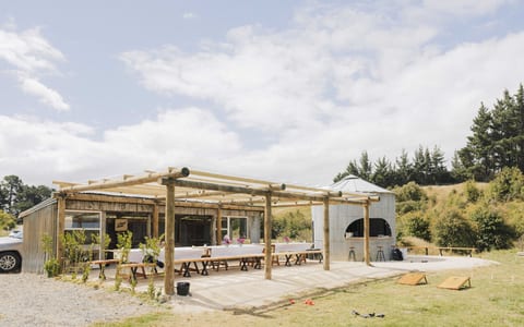 Open timber-frame outdoor shelter with long trestle tables, corrugated iron bar structure beside it, paddock setting