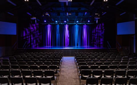 Large performing arts auditorium with stage, curtains lit in purple and blue, and rows of seats