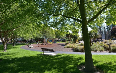 Park with trees, lawn and playground equipment visible, bench in foreground