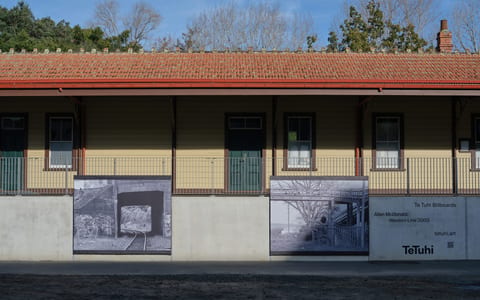 Single-storey yellow weatherboard building with red tile roof, large billboard artworks on base