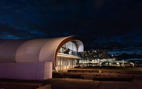 White curved barrel-vault building at night on a waterfront, city lights behind