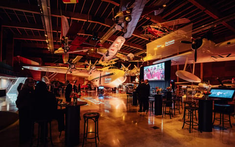 Event space inside a museum hall with vintage aircraft suspended overhead and cocktail tables