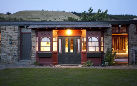 Stone stable building at dusk with lit interiors through arched windows opening onto a lawn