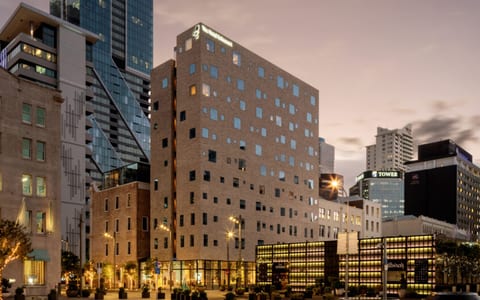 Pale brick multi-storey building with irregular window openings, city skyline at dusk