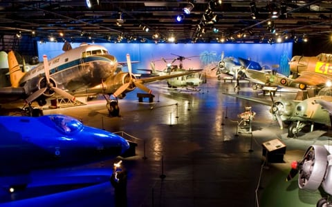 Large darkened museum hall with multiple historic aircraft on display under coloured spotlights