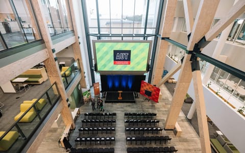 Large atrium event space with timber columns, rows of chairs, and a raised screen displaying a logo
