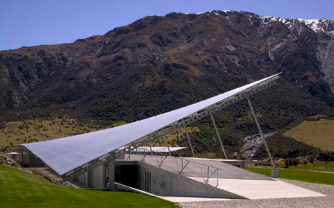 Low angular metal-roofed building embedded in hillside with mountain backdrop and open gravel forecourt