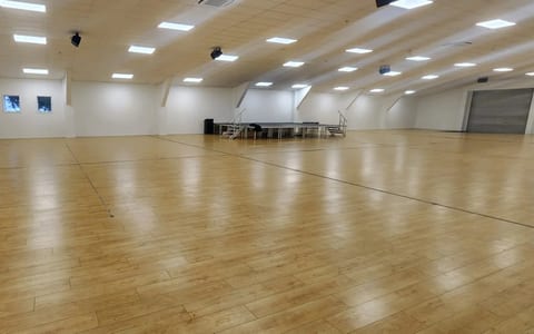 Large empty hall with timber floor and raised platform at the far end, lit by overhead panels