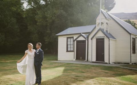 A couple in wedding attire on a lawn beside a small white weatherboard chapel, framed by tall trees with hills behind