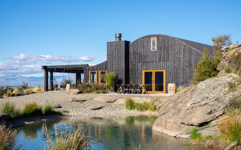 Dark timber barn with curved roof beside a pond, rocky landscape, open mountain backdrop