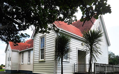 White weatherboard hall with red corrugated roof and cabbage trees at entrance