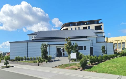 Single-storey white weatherboard building with dark trim, sloped metal roof, suburban setting