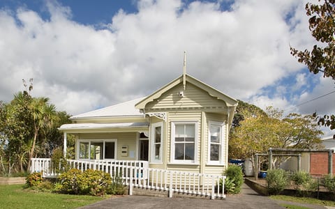 Cream weatherboard villa with verandah, bay window, white picket fence, and lawn, sunny suburban setting