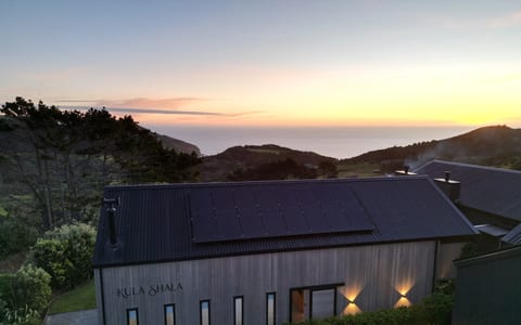Timber-clad building with a dark metal roof and solar panels, viewed from above at sunset with sea behind