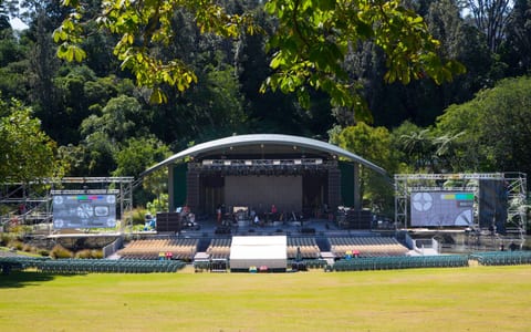 Outdoor amphitheatre with covered stage, seating, and large screens in parkland