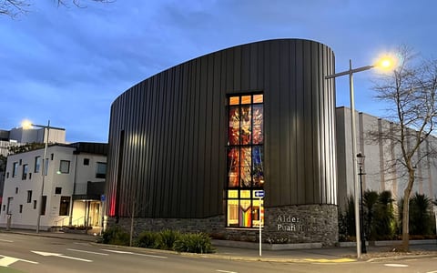 Contemporary curved dark-clad church building at dusk with stained glass feature window