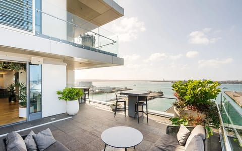 Rooftop terrace with grey sofas and planters, glass balustrade, and harbour and cityscape view