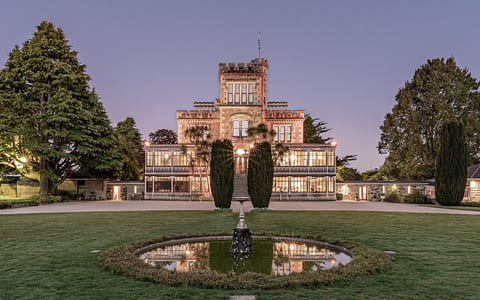 A lit stone castle with a tower and glasshouse wing at dusk, framed by tall trees and a formal lawn with a circular fountain pool