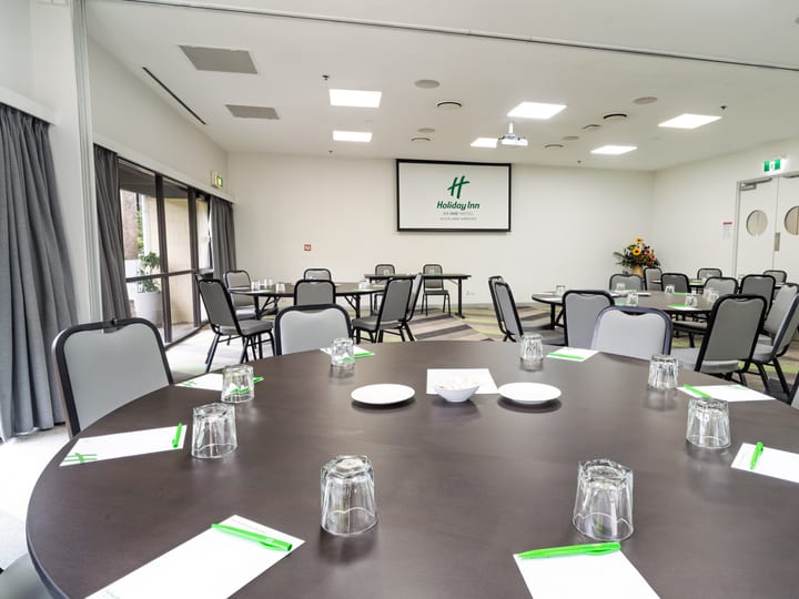 Plain white function room with round tables, mixed seating, and a projector screen at one end