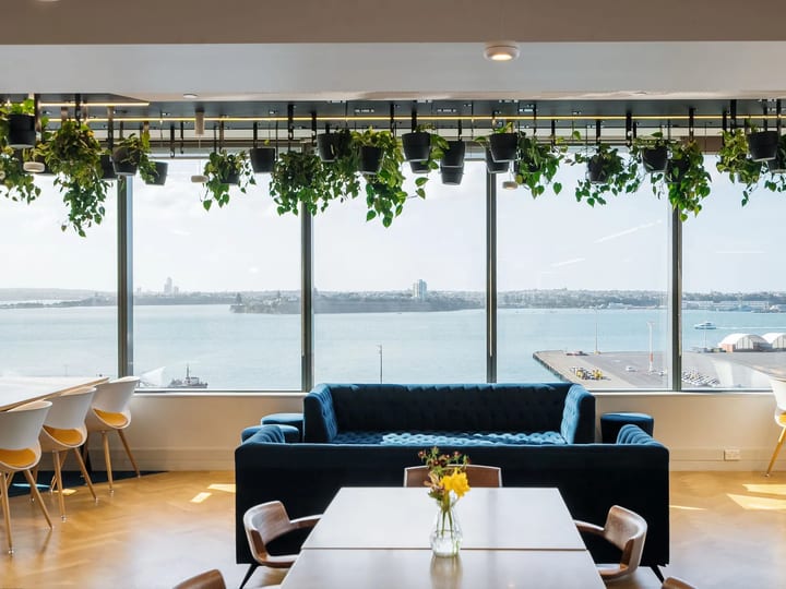 Upper-floor lounge with blue velvet sofa, hanging potted plants, and panoramic harbour view through full-height windows