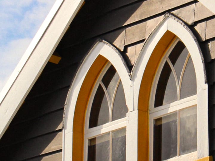 Grey weatherboard gable end with arched Gothic-style windows and yellow surrounds against blue sky