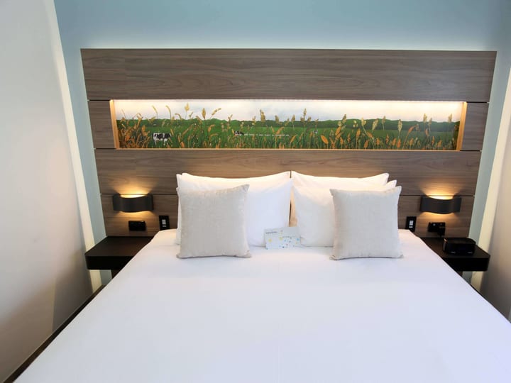 Hotel bed with timber headboard, backlit landscape artwork panel, and flanking bedside lamps