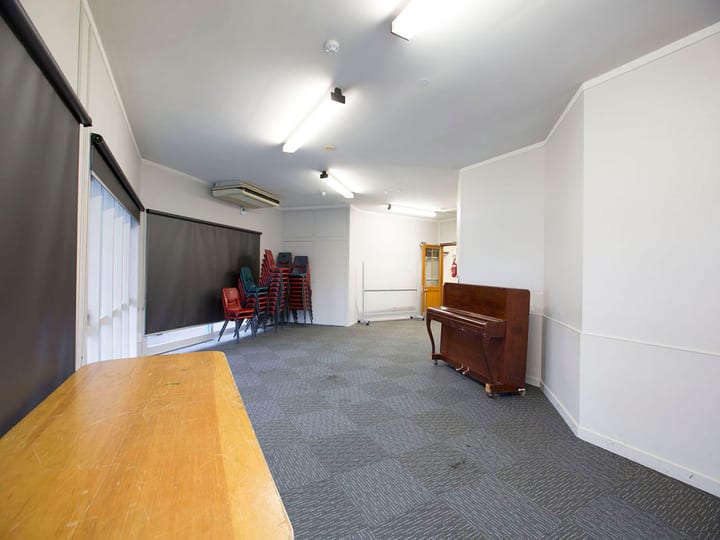 Carpeted meeting room with stacked chairs, upright piano, and roller blinds