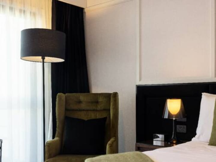 Hotel room with double bed, dark bedhead, green upholstered armchair, and sheer curtains
