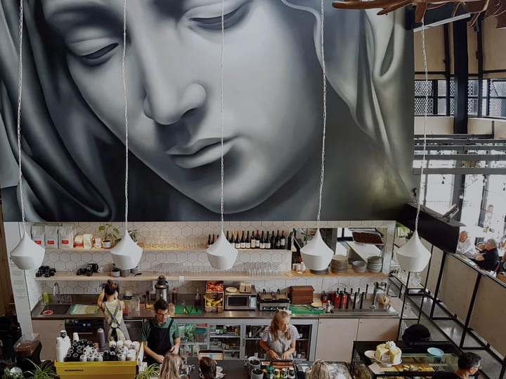 Double-height cafe interior with large mural on rear wall, bar counter, hanging pendant lights, industrial shelving