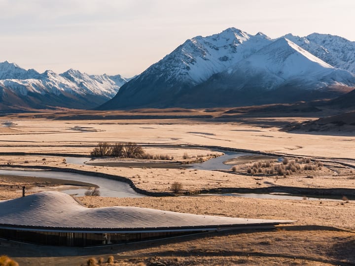 Low-profile curved building on a flat river plain with snow-capped mountains behind