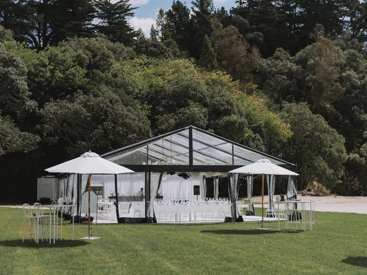 Glass and steel clear-span marquee on lawn with white chairs inside, trees and blue sky behind
