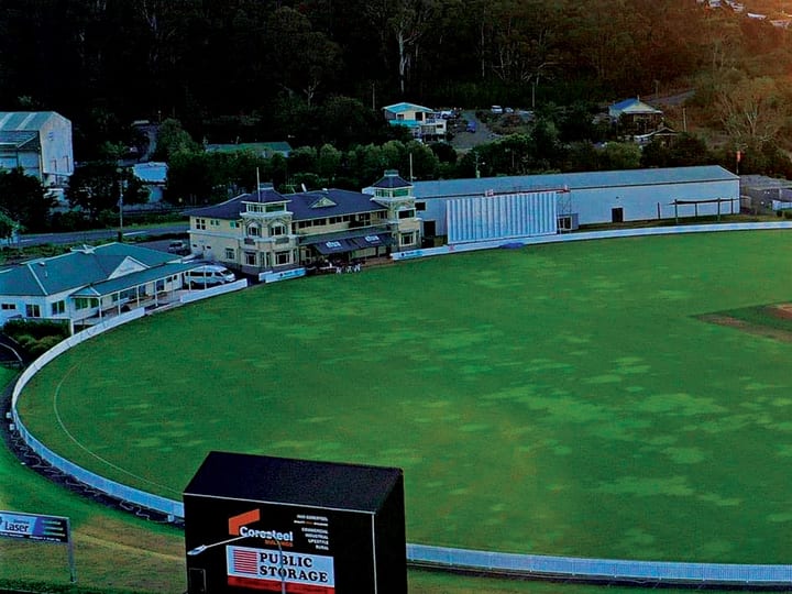 Cricket oval at dusk with a scoreboard in the foreground and surrounding hills