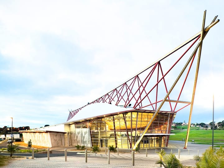 Event building exterior by day with angled red steel lattice canopy, glazed lower volume, and sculpture nearby