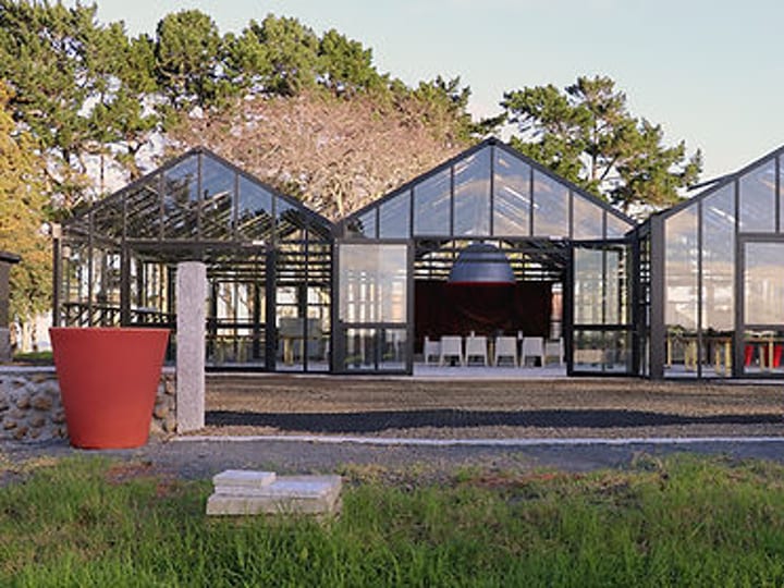 Black steel glasshouse exterior with white chairs visible inside, large planters flanking entrance