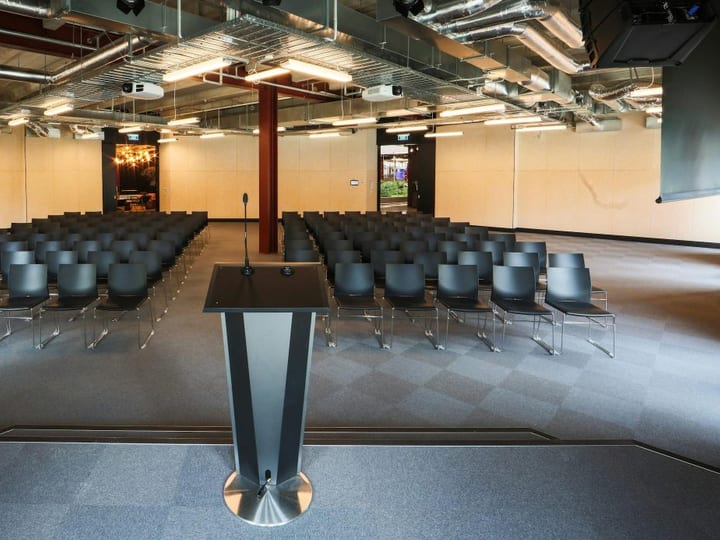 Conference hall with rows of black chairs facing a lectern, dual projection screens, and exposed ducting