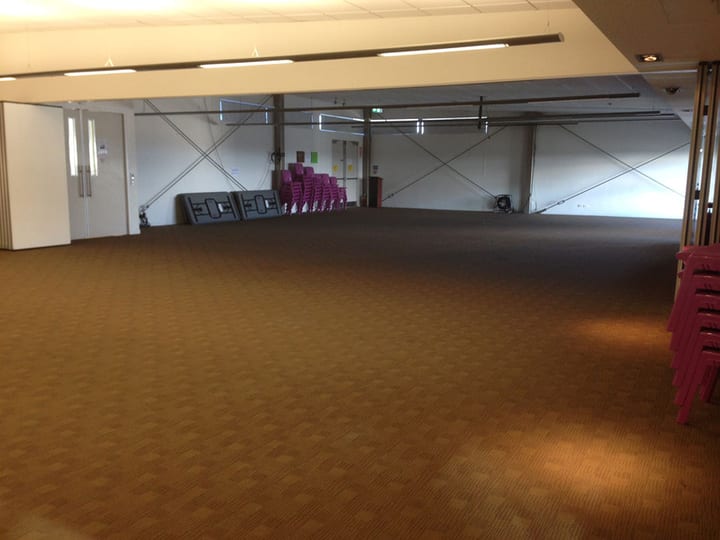 Large carpeted function hall with bracing cables, stacked chairs and pink chairs