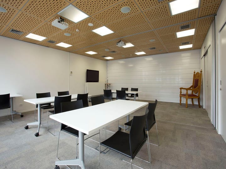 Meeting room with perforated timber ceiling, white tables, dark chairs, wall-mounted screen, and ornate timber chair in corner