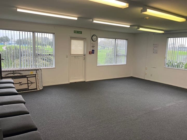 Plain carpeted room with fire exit door, two windows and folded cushions along one wall