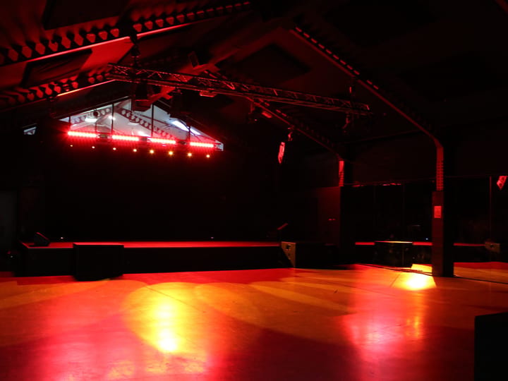 Dark warehouse interior with red stage lighting, exposed truss and open concrete floor