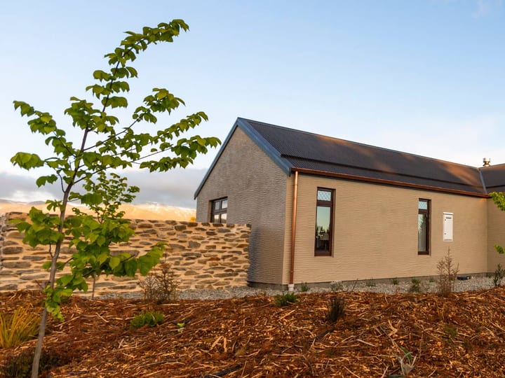 Cream render cottage with dark metal roof and timber gable porch, young plantings at dusk