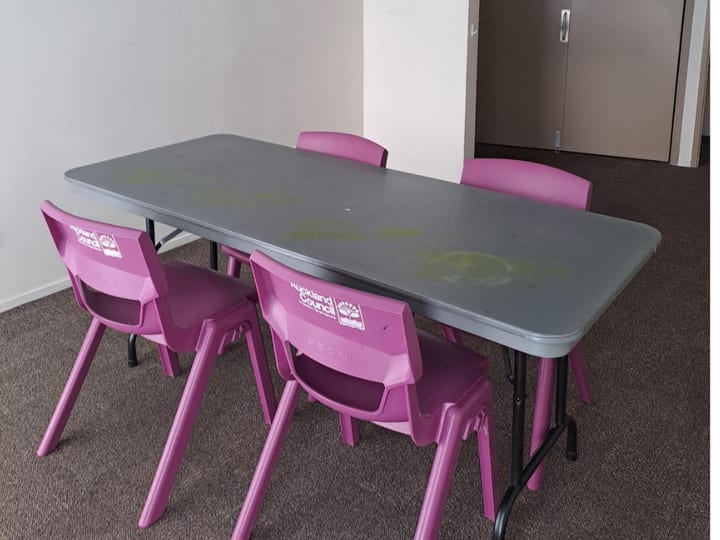 Small carpeted room with pink plastic chairs around a grey trestle table